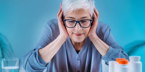 Person in hellblauer Kleidung sitzt an einem Tisch, hält beide Hände an den Kopf, vor sich ein Glas Wasser und mehrere Medikamentenbehälter.